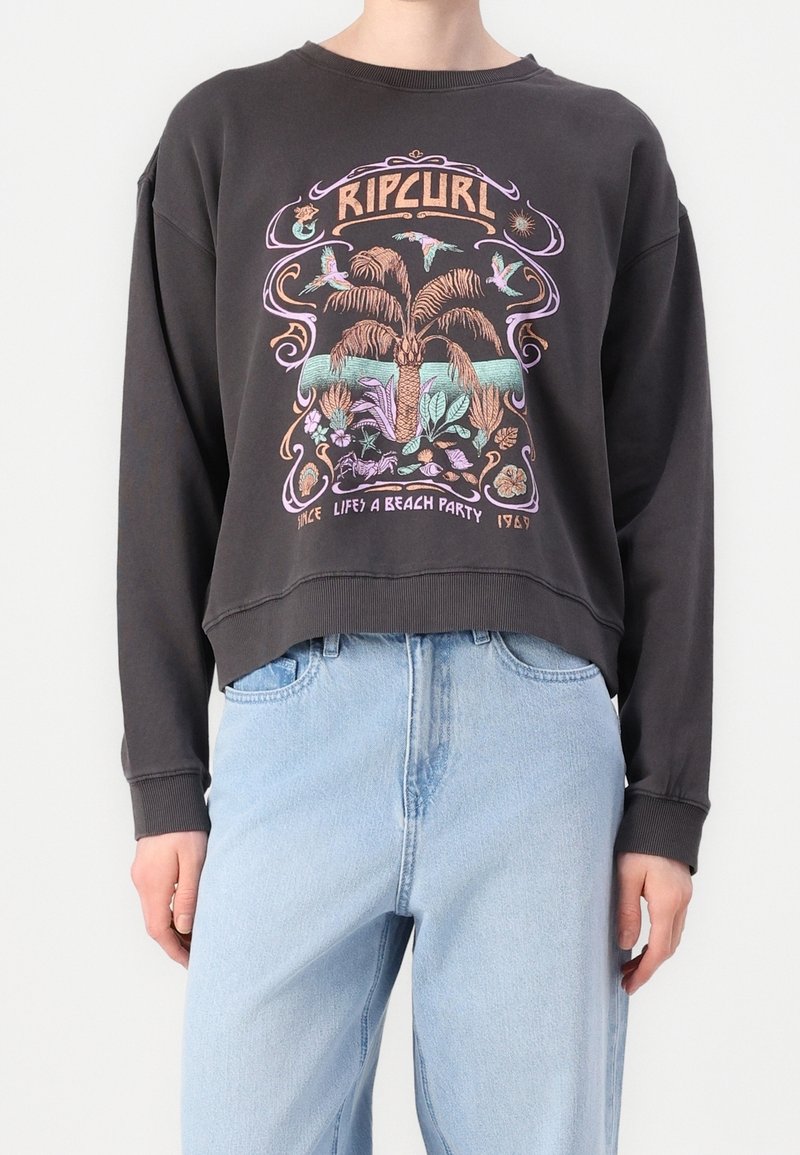 Sweat-shirt noir avec imprimé tropical coloré, palmier, oiseaux et texte "Rip Curl", associé à un jean bleu clair.