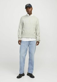 Suéter de punto gris claro con cuello redondo, combinado con una camisa blanca de cuello, pantalones vaqueros azules y zapatos negros.
