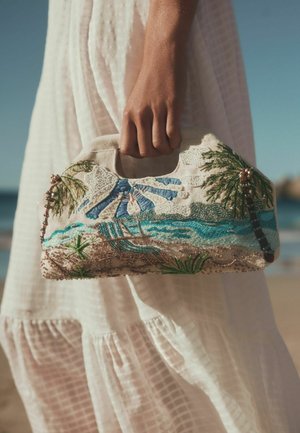 Roka tur nelielu auduma rokassomiņu ar izšūtu pludmales ainavu, kurā ir palmas, okeāna viļņi un atpūtas krēsli, baltas kleitas priekšā.