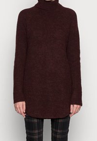 Suéter de punto burdeos con cuello alto, mangas largas y dobladillo redondeado, que muestra una textura suave y un ajuste holgado. Combinado con pantalones a cuadros.