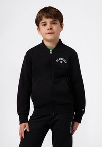 Niño pequeño vestido con chaqueta de cremallera negra de Champion y pantalones de chándal negros a juego, de pie contra un fondo blanco.