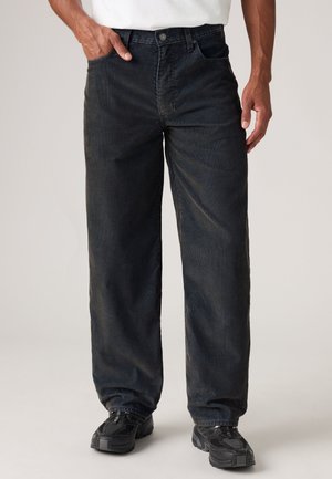 Homme portant un pantalon en velours côtelé noir ample et des baskets noires, debout avec une main dans la poche devant un fond uni.