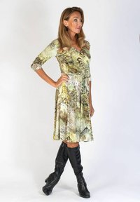 Nainen seisoo käsi lantiolla, yllä vihreäkuvioinen polveen asti ulottuva mekko ja mustat polvisukat, yksinkertaista vaaleaa taustaa vasten.