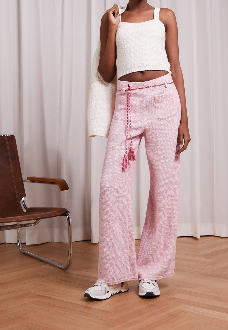 Personne portant un débardeur tricoté blanc, tenant un pull assorti, un pantalon large rose avec une ceinture à franges et des baskets blanches, debout à l'intérieur.