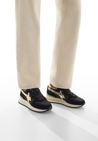 Sneaker nere e oro con tomaia in suede e mesh, dotate di un grande logo a stella e una suola intermedia bianca, abbinate a pantaloni color crema.