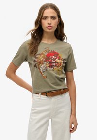Camiseta de algodón verde oliva con gráfico de un tigre, sol rojo y acentos dorados. Mangas cortas, cuello redondo, combinada con pantalones blancos y un cinturón marrón.