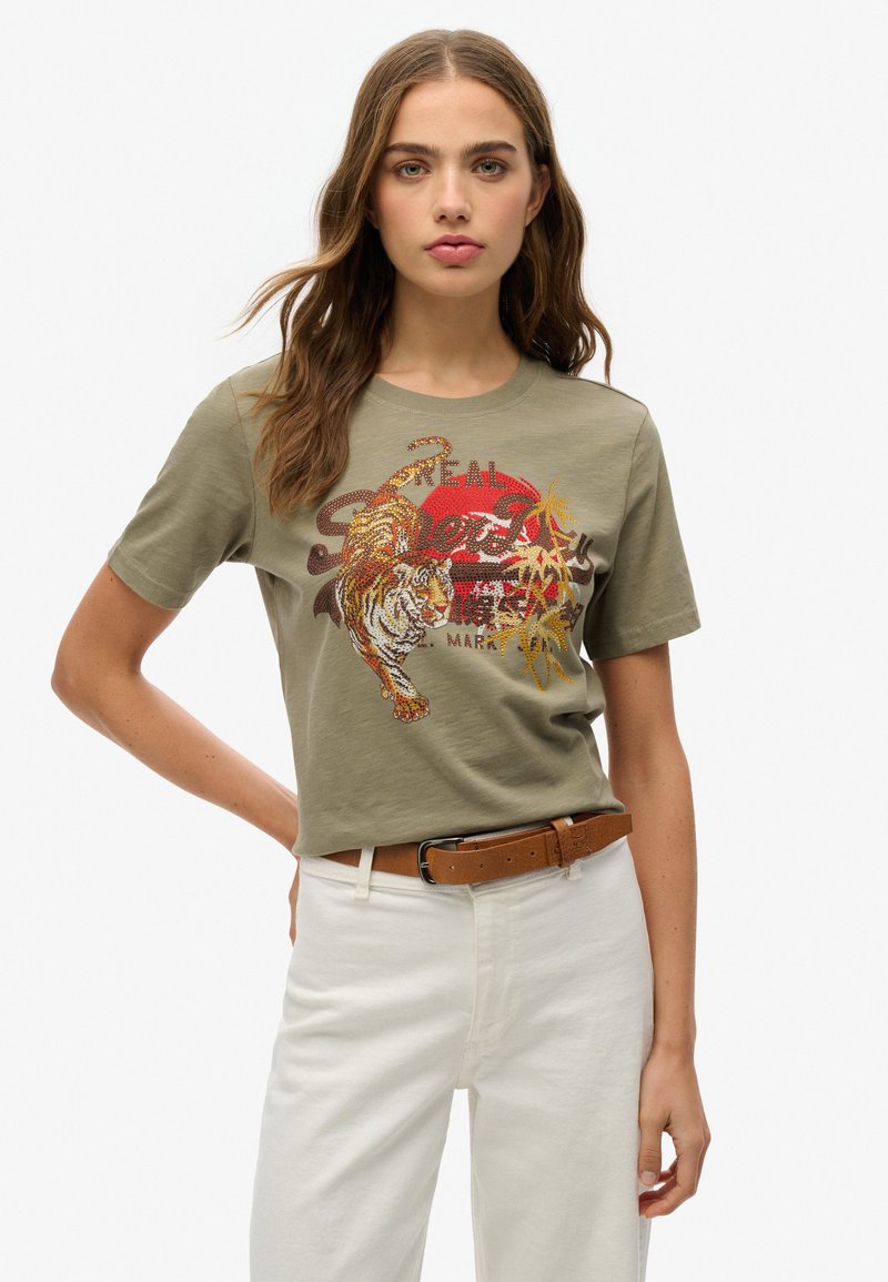 Camiseta de algodón verde oliva con gráfico de un tigre, sol rojo y acentos dorados. Mangas cortas, cuello redondo, combinada con pantalones blancos y un cinturón marrón.