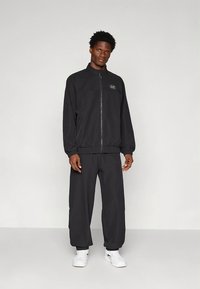 Svart treningsdress med glidelåsjakke med lomme og elastiske mansjetter, kombinert med løstsittende bukser og hvite joggesko.