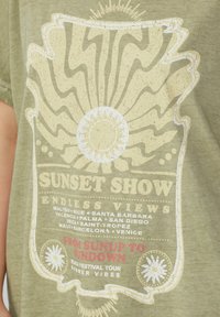 Olivgrünes T-Shirt mit einem Sonnenmotiv in Beige und Weiß, mit festivalthematischen Texten in Rot und Weiß am unteren Rand.