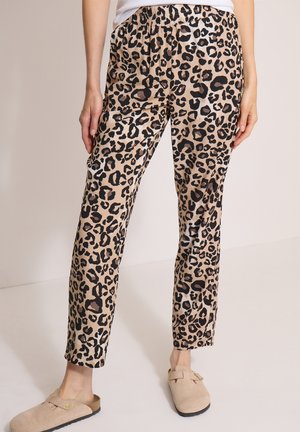 Person iført løse leopardmønstrede bukser, hvid skjorte og beige hjemmesko, står indendørs mod en lys ensfarvet baggrund.