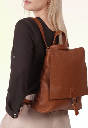 Vrouw met lang blond haar, gekleed in een zwarte blouse en lichte rok, draagt een gestructureerde bruine leren rugzak met gesp sluiting.