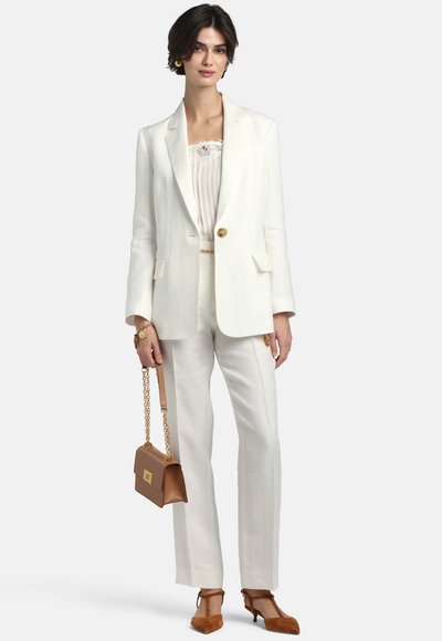 Fehér blézert és nadrágot viselő nő, barna láncos pántú kézitáskát tart, barna orrfazonú magassarkút és arany ékszereket visel.