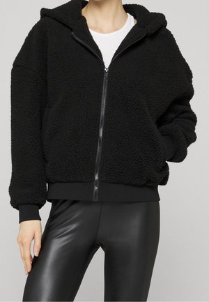 Osoba nosząca czarną polową bluzę z kapturem zapinaną na suwak z kieszeniami oraz czarne legginsy ze sztucznej skóry, pod bluzą widoczna biała koszulka.