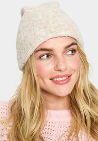 Beannie lavorato a maglia color crema con superficie testurizzata, cima arrotondata e bordo arrotolato. Indossato con un maglione rosa chiaro con un motivo in pizzo.
