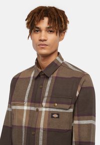Camicia a quadrati marrone realizzata in un tessuto morbido, con linee bianche e viola a contrasto, una chiusura frontale con bottoni e una tasca sul petto con un logo.