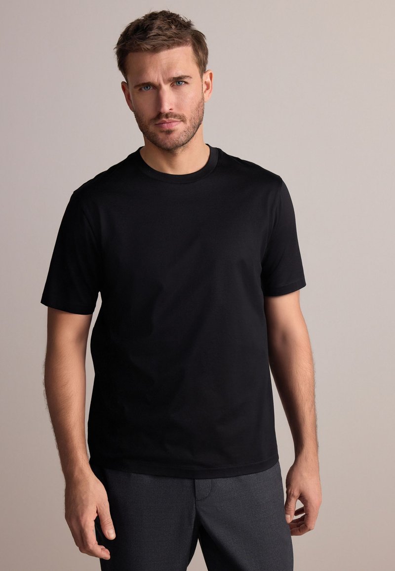 Homme aux cheveux courts et bruns avec une barbe, portant un t-shirt noir uni à manches courtes et un pantalon gris foncé, debout devant un fond uni.