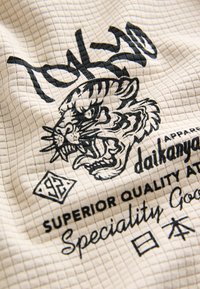 Tessuto intrecciato con motivo a griglia, colore crema. Presenta una grafica nera di una tigre e testo con le parole "Tokyo" e "Daikanyama".