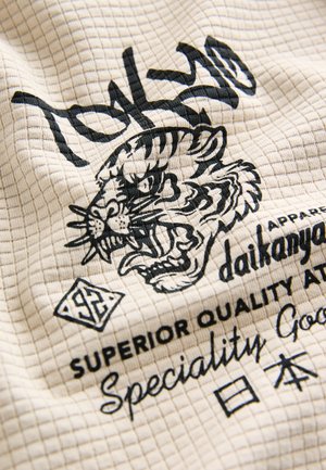 Kootud tekstiil ruudulise mustriga, kreemjas värv. Kujutisena on must tiigri graafika ning tekst "Tokyo" ja "Daikanyama".