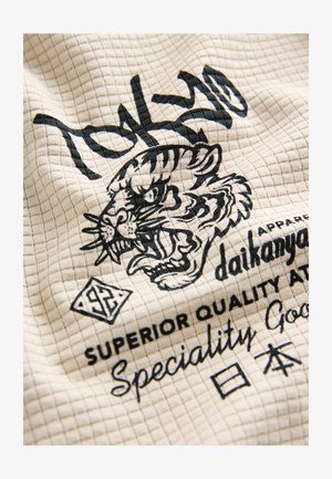 Tkany materiał z wzorem w kratkę, kolor kremowy. Zawiera czarną grafikę tygrysa oraz tekst "Tokyo" i "Daikanyama".