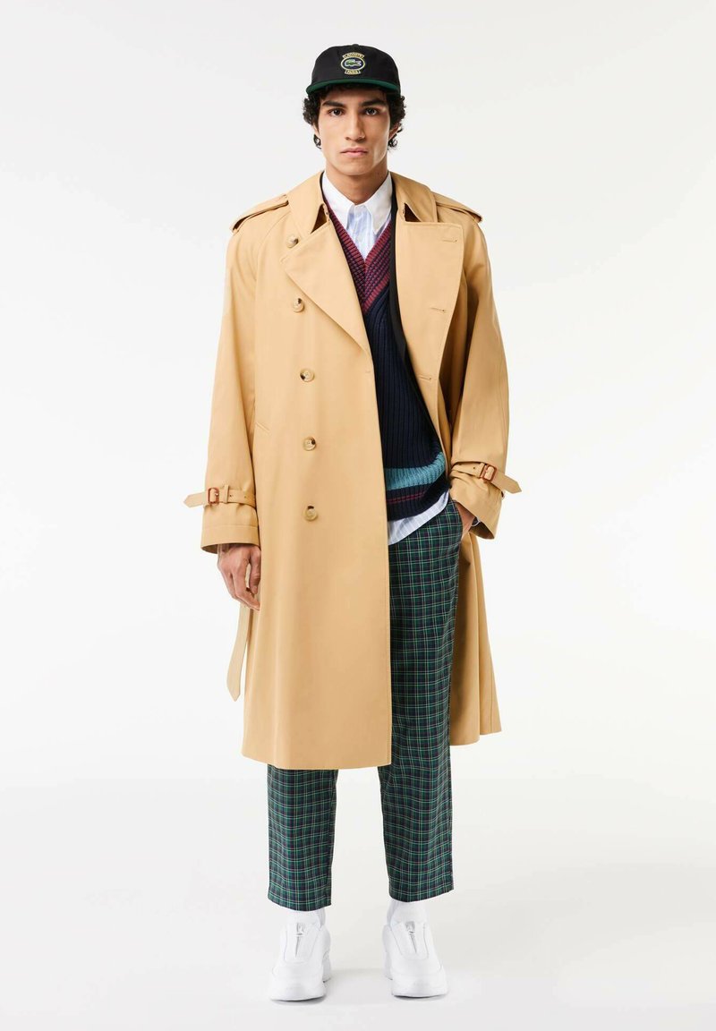 Manteau trench beige à boutons double boutonnage, ceinturé à la taille, superposé à un pull bleu marine, associé à un pantalon à carreaux verts et des baskets blanches.