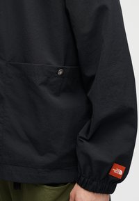 Μανίκι μαύρου μπουφάν με ελαστική μανσέτα και κόκκινη ορθογώνια ετικέτα λογότυπου, τσέπη με λεπτομέρεια μεταλλικού κουμπιού κλικ, φορεμένο με παντελόνια ελιάς πράσινου χρώματος.