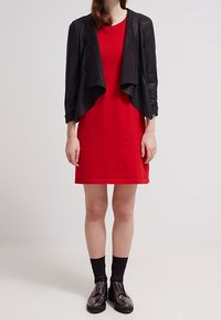 Giacca nera corta con un fronte drappeggiato sopra un vestito rosso. Tessuto liscio, vestito lungo fino al ginocchio, calze nere e scarpe nere lucide.