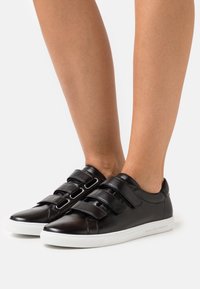 Baskets en cuir noir avec deux bandes Velcro, bout rond et semelle en caoutchouc blanc. Texture lisse avec des détails de design minimalistes.