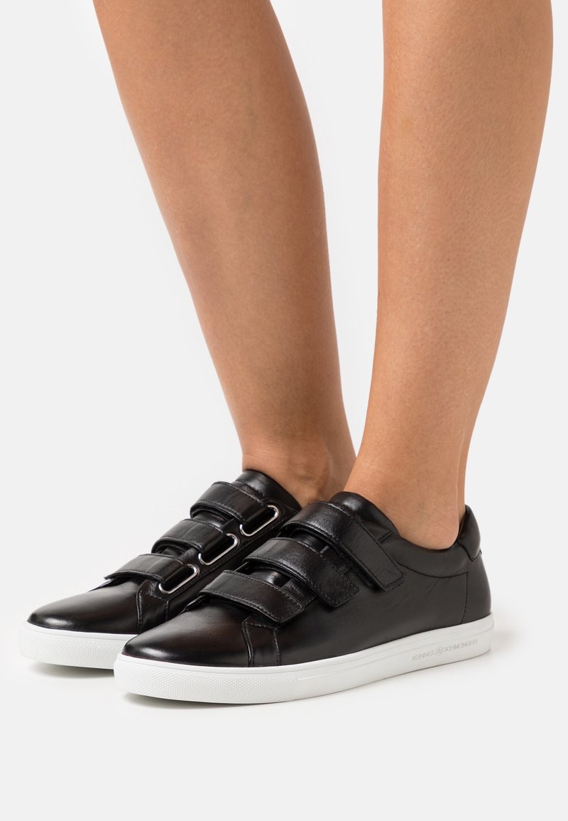 Baskets en cuir noir avec deux bandes Velcro, bout rond et semelle en caoutchouc blanc. Texture lisse avec des détails de design minimalistes.