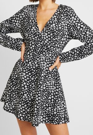 Kobieta w czarnej sukience w białe kwiaty, z długimi rękawami i dekoltem w serek, pozująca z rękami na biodrach na jednolitym tle.