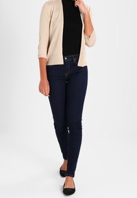 Beige gebreide cardigan, zwarte aansluitende top, donkerblauwe skinny jeans en zwarte platte schoenen. De cardigan heeft driekwart mouwen en een open voorkant.