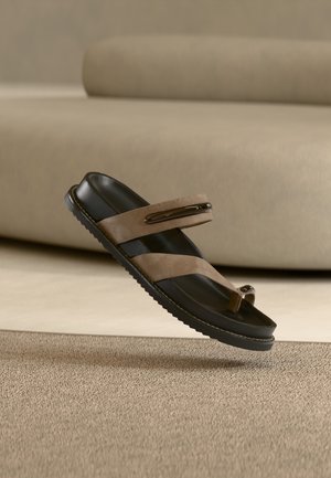 Brown and black open-toe sandal with two straps and toe loop, floating above a textured beige carpet and beige couch background.