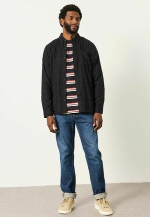 Man wearing black button-up shirt, striped t-shirt, blue jeans rolled at cuffs, and beige sneakers standing against plain background.