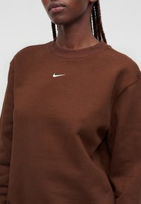 Άτομο που φοράει καφέ φούτερ Nike με λογότυπο Swoosh σε λευκό, με ορατά κοτσίδια, σε απλό ανοιχτό φόντο.