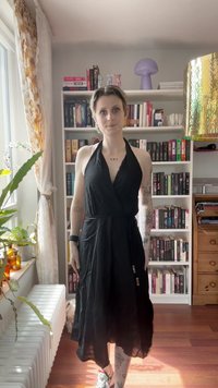 Vestido preto halter com um design envolvente, apresentando uma silhueta drapeada e um tecido suave. Detalhes notáveis incluem uma fita na cintura.