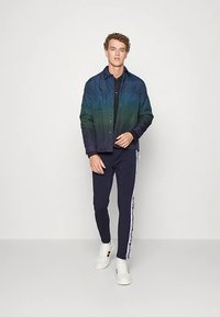 Ung mann iført en gradient blå-grønn skjortejakke, svart skjorte, marineblå joggebukser med hvite side-striper, og hvite sneakers som står mot en ensfarget bakgrunn.