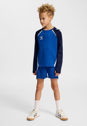 Junges Kind mit lockigem Haar, das blaue und marineblaue Sportkleidung, weiße Socken und weiße Sportschuhe trägt, steht und schaut zur Seite.