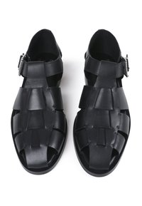 Sandales noires avec un design tressé, fabriquées en matériau lisse et mat. Équipées d'une sangle réglable à la cheville et d'un bout ouvert. Semelle plate.