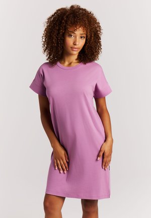 Frau mit lockigem Haar, die ein schlichtes kurzärmeliges fliederfarbenes Kleid trägt und mit entspannten Händen in Hüfthöhe vor einem hellen Hintergrund steht.