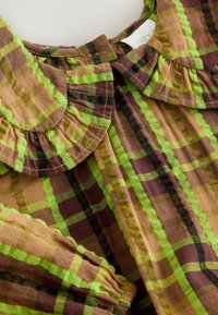 Braune und grüne Karobluse mit Rüschenspul. Der Stoff ist aus leichtem Baumwollmaterial und zeigt ein kariertes Muster mit kontrastierenden Farben.