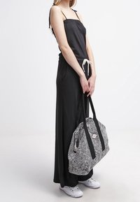 Sort ærmeløs jumpsuit med snoretræk i taljen; modellen holder en sort og hvid mønstret tote bag; hvide sneakers fuldender looket.