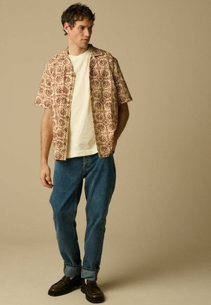 Homme portant une chemise à manches courtes beige à motifs par-dessus un t-shirt blanc, jean bleu retroussé et mocassins marron foncé, debout devant un fond beige uni.