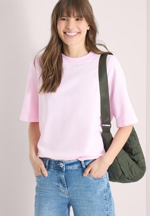 Femme souriante portant un t-shirt rose clair à manches courtes, un jean bleu et un sac à bandoulière matelassé vert foncé, les mains dans les poches.
