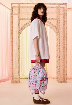 Noor naine valges särgis ja punases seelikus hoiab lilla lillemustriga seljakotti, seisab pastelses toonis ruumis läbipaistvate kardinatega ja kaarega.