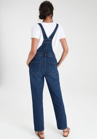 Salopette en denim avec bretelles réglables, coupe décontractée, deux poches arrière et teinte bleu moyen, associée à un t-shirt blanc.