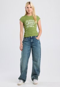 Green fitted t-shirt featuring white graphic text; paired with loose-fit blue denim jeans; model stands with hands on hips, wearing white sneakers.