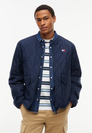 Ung mand iført en mørkeblå quiltet jakke over en blå denimskjorte og en stribet T-shirt, med beige cargobukser, stående med hænderne i lommerne.