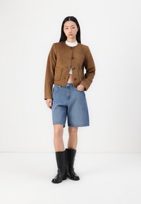 Brun suedet cropped jakke med knaplukning, to lommer, parret med denim shorts og sorte knæhøje støvler. Hvid top nedenunder.