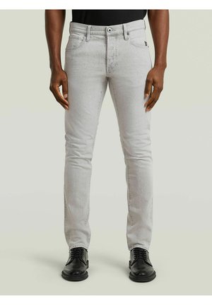 Homme portant un jean slim gris clair et des chaussures habillées noires, debout devant un fond clair uni.