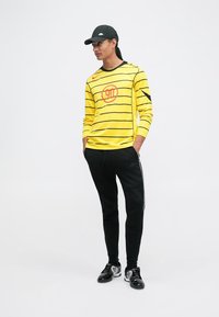 Geel shirt met lange mouwen en zwarte strepen, gecombineerd met zwarte trainingsbroek met een witte zijsstrijp. Zwarte pet en sneakers.