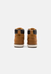 Botas altas de cuero marrón claro con un cuello acolchado negro, suela blanca y costuras visibles. Presenta una cinta en la parte trasera y un pequeño detalle de logo.